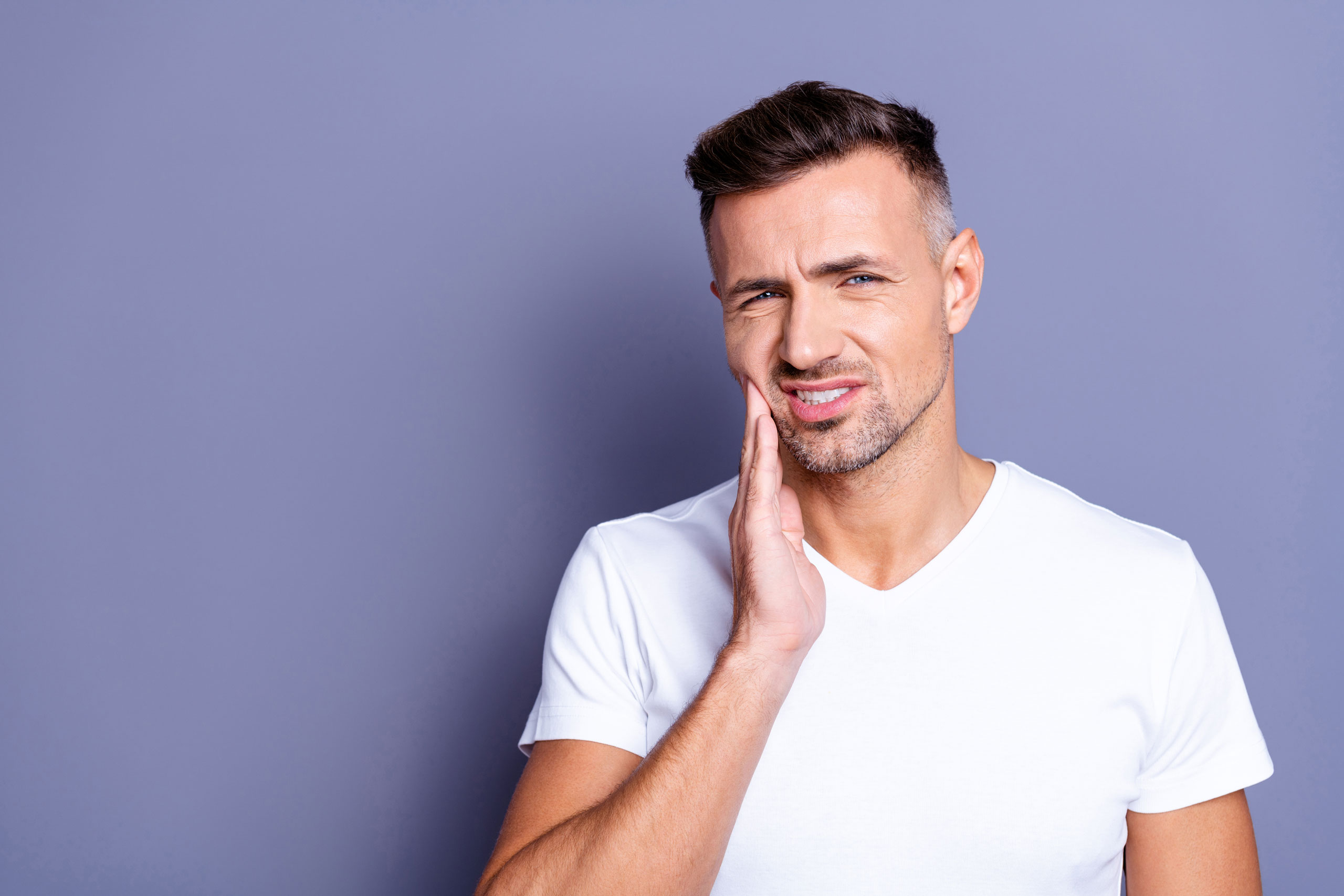 Young man with short brown hair and trimmed beard, wearing a white t-shirt against a purple wall. He is wincing in pain and holding his face with his right hand due to a toothache.