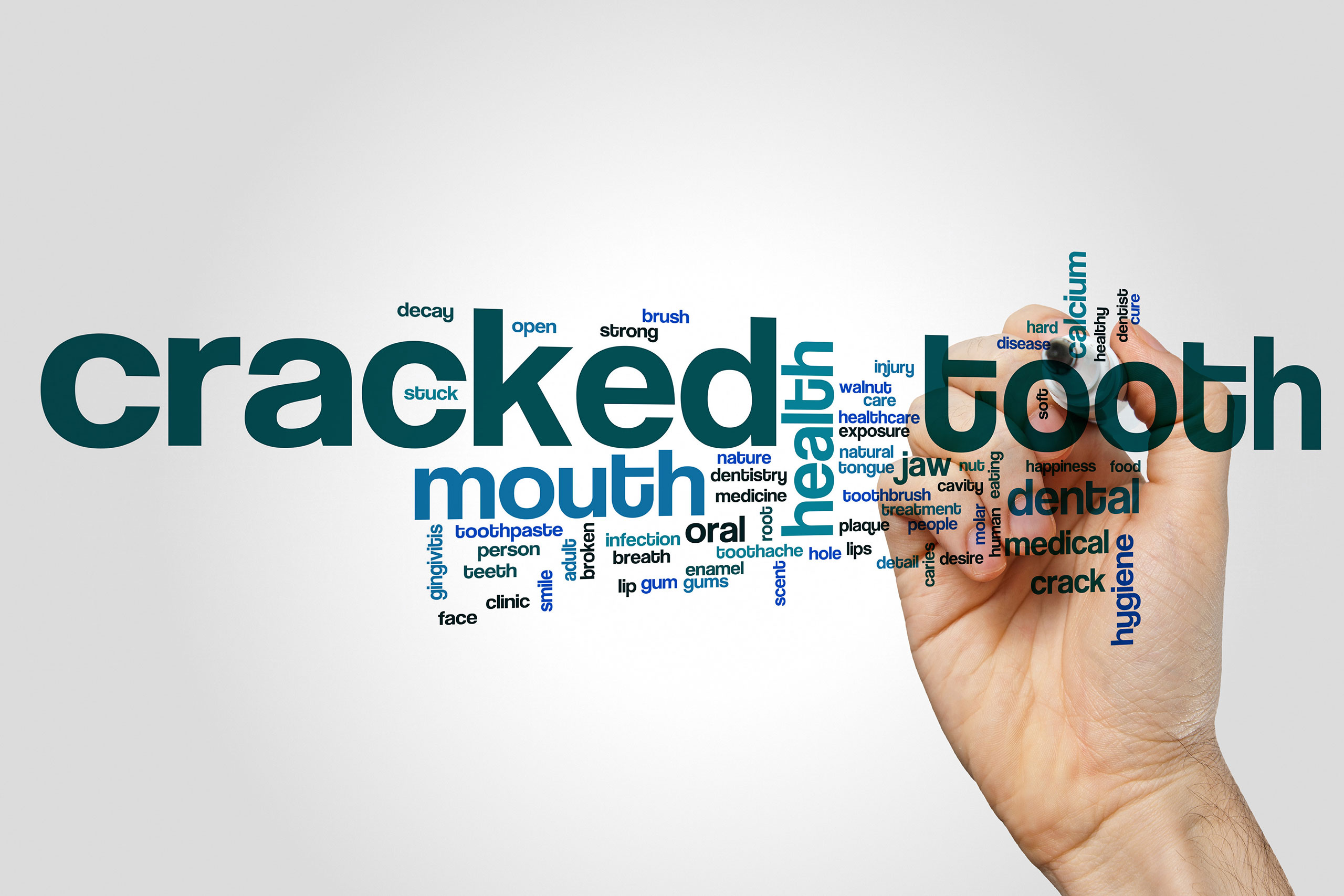 A word cloud focused on the terms "cracked tooth," with related words in various sizes, such as "mouth," "health," "dental," "jaw," and "hygiene." The text appears in shades of blue and black, with larger words indicating higher relevance. A hand on the right side of the image is holding a marker as if drawing or writing the words. The background is a plain white, keeping the focus on the text.