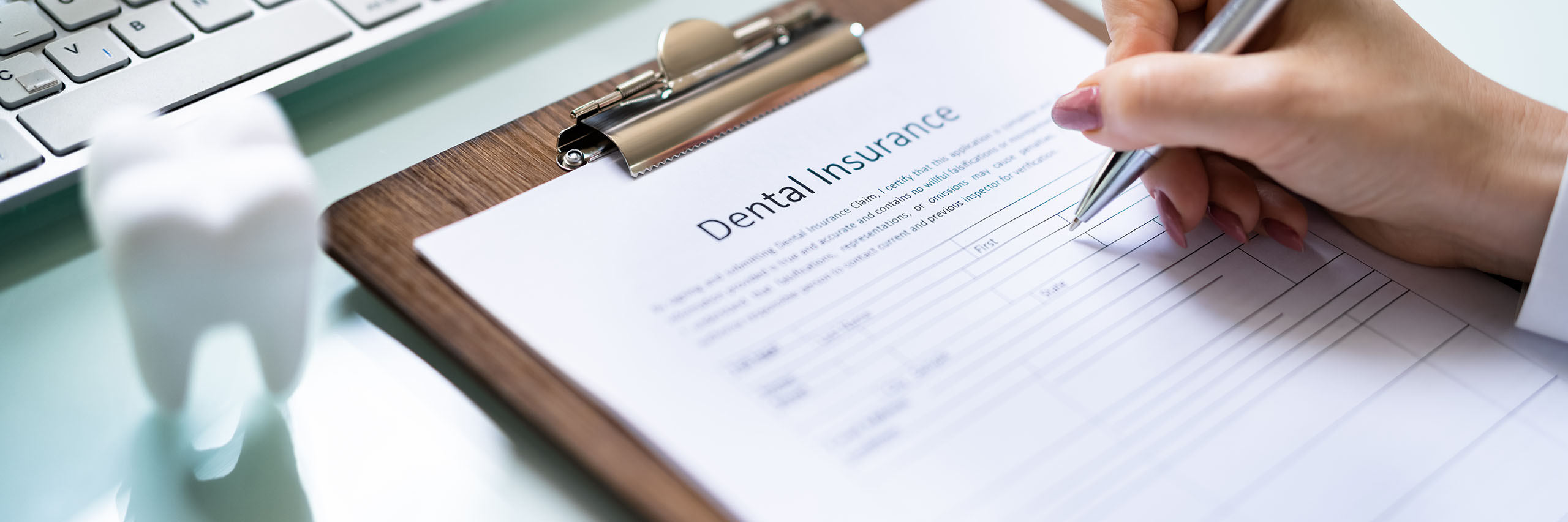 A person filling out a dental insurance form on a clipboard, with a pen in hand. The form is titled "Dental Insurance" at the top. A computer keyboard and a small model of a tooth are visible in the background on a glass desk, suggesting a dental or office setting.