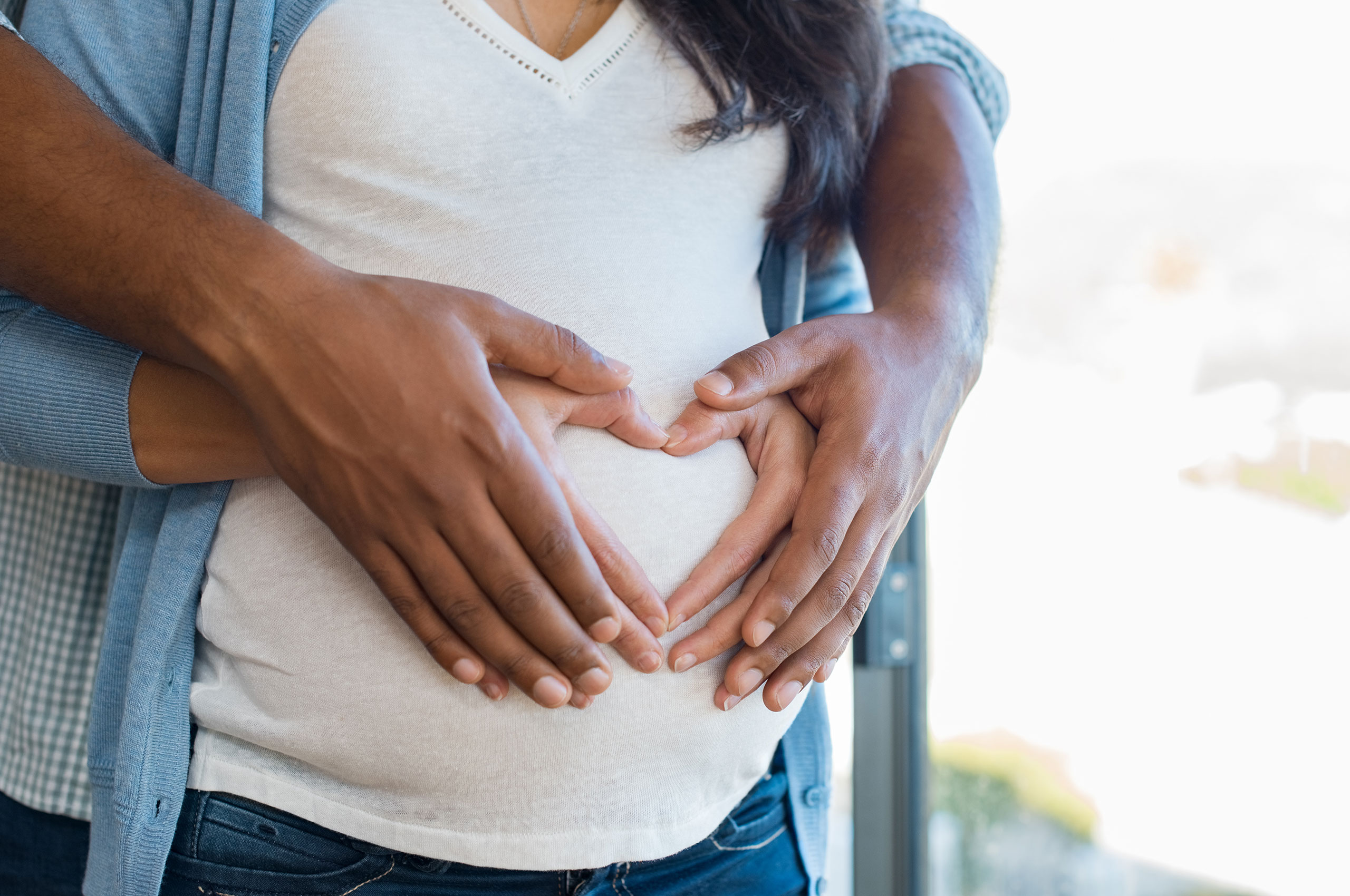 Close-up of a pregnant woman's belly with her hands and her partner's hands gently placed over it. Their fingers form a heart shape, symbolizing love and anticipation for the baby. The woman wears a white top and a blue cardigan, while the partner embraces her from behind. The background is softly blurred with natural light coming through a window.
