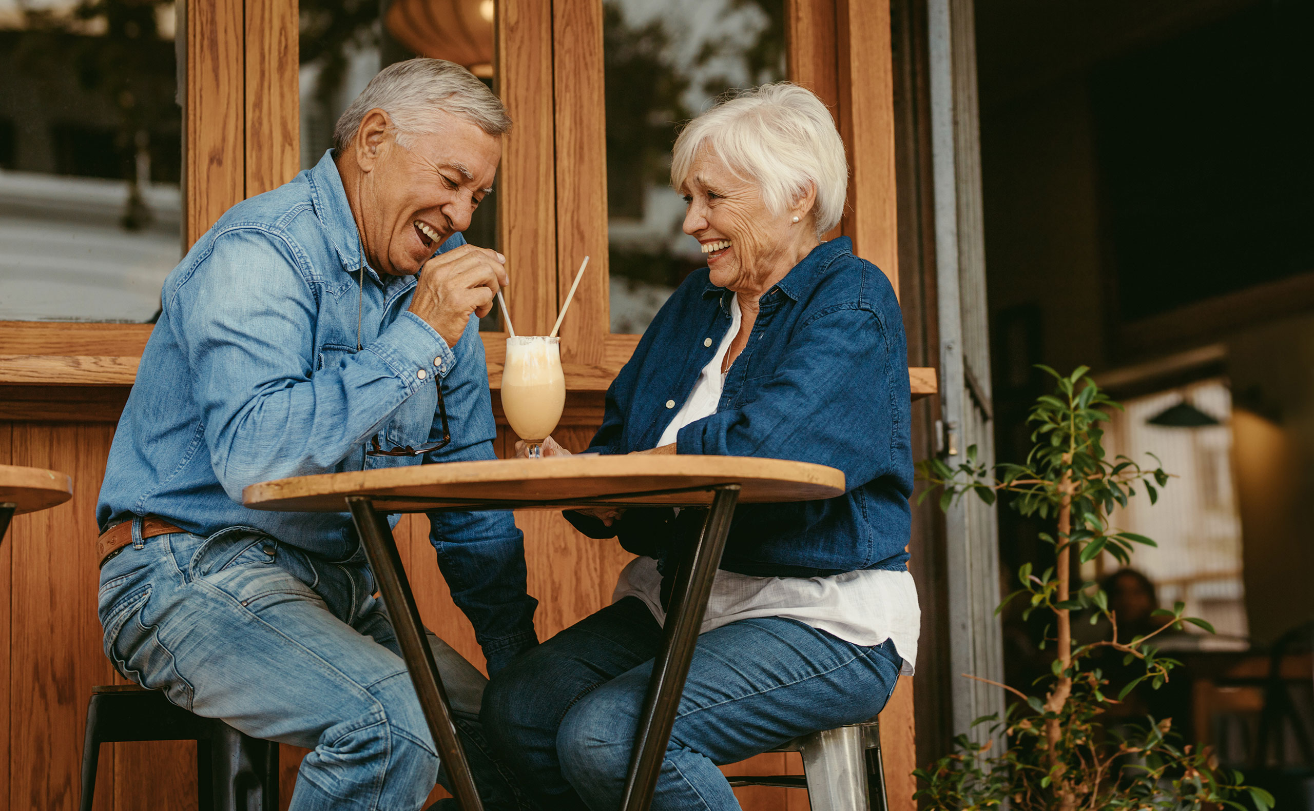 Elderly couple sitting at an outdoor café table, smiling and laughing while sharing a milkshake with two straws. Both are wearing denim outfits and appear to be enjoying each other’s company. The café has wooden walls and large windows, with a small potted plant nearby.