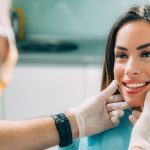 Smiling woman at a dental clinic being examined by a dentist wearing gloves; the dentist gently holds her chin while assessing her teeth.