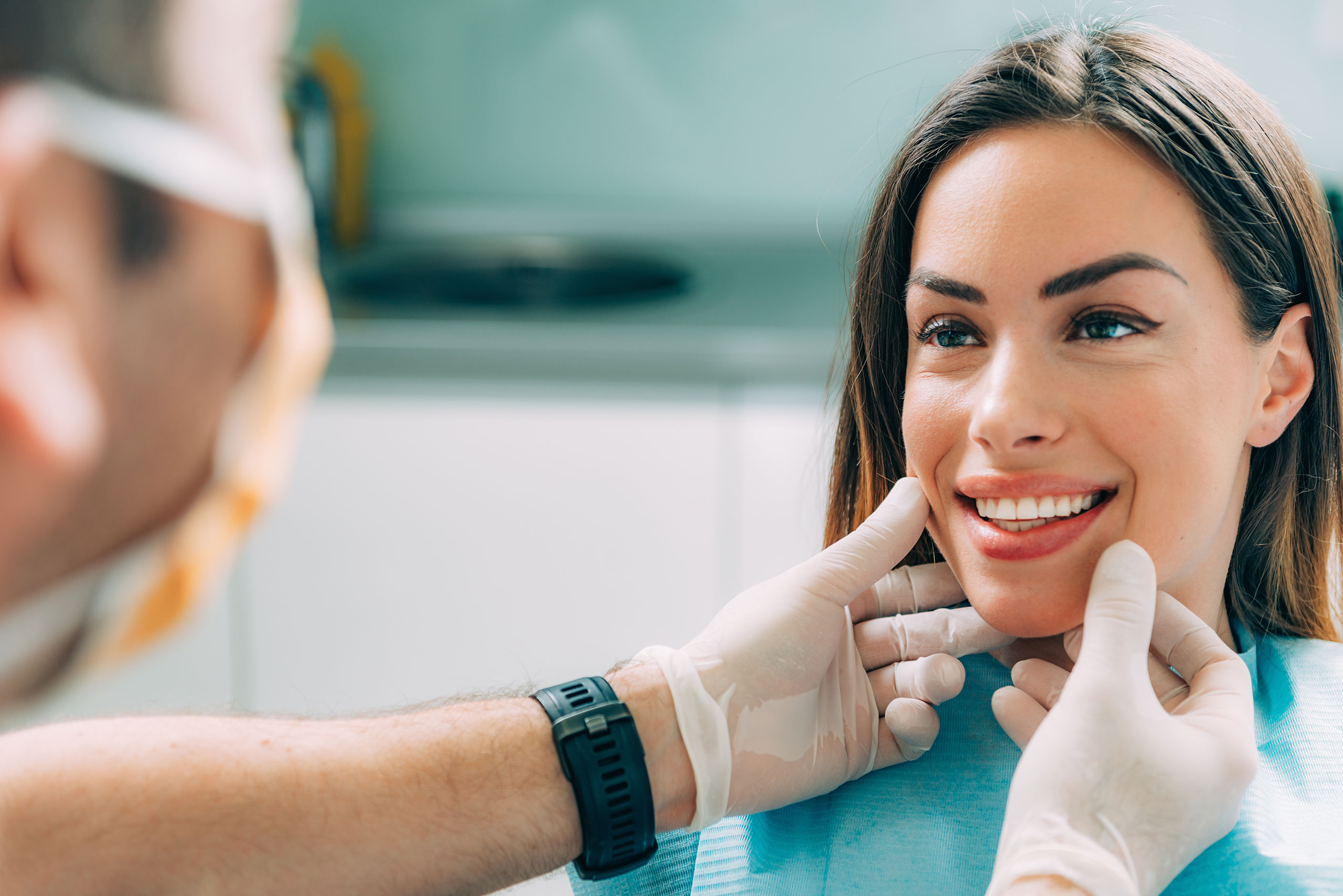 Smiling woman at a dental clinic being examined by a dentist wearing gloves; the dentist gently holds her chin while assessing her teeth.