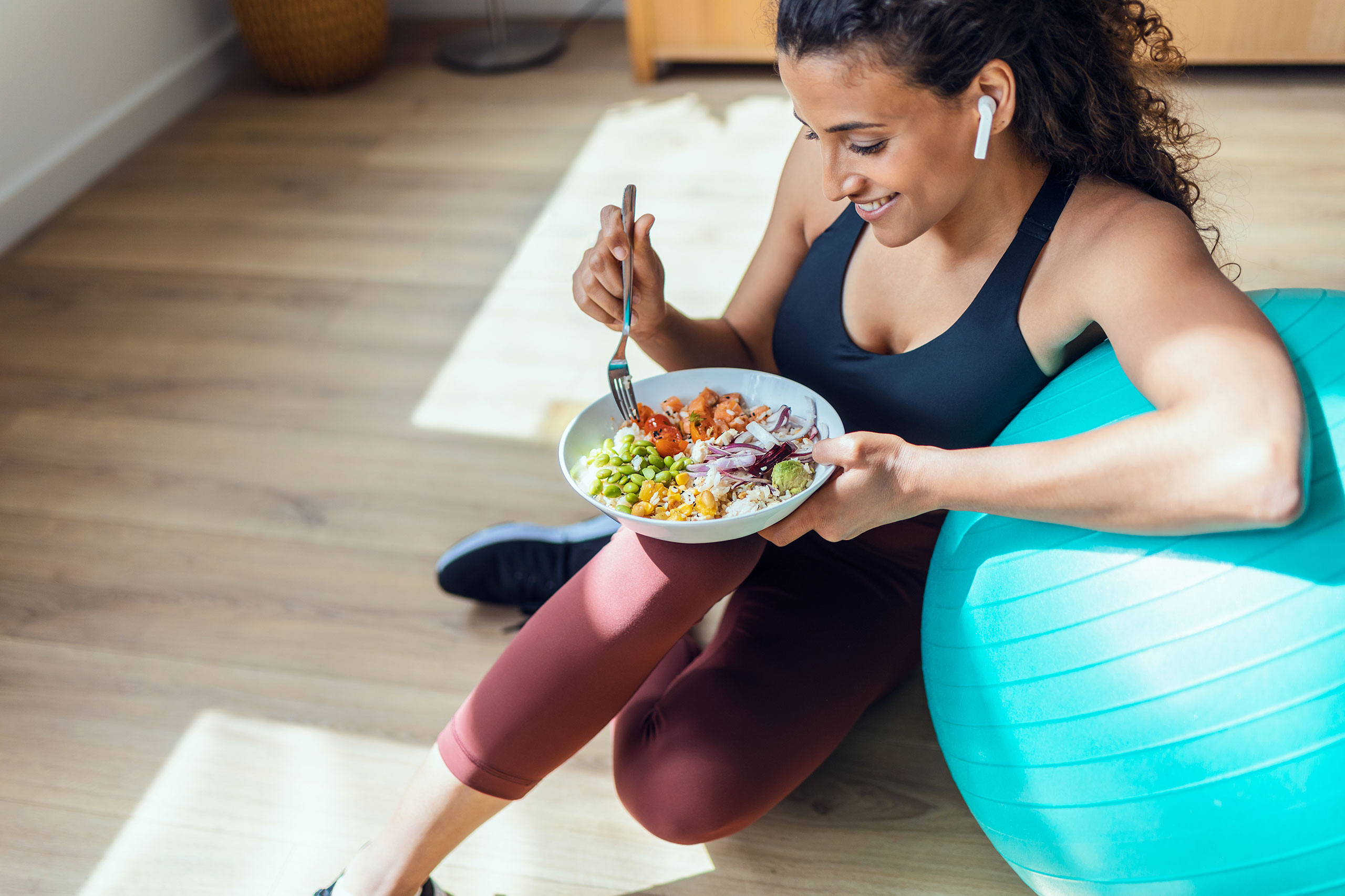 Woman in athletic wear sitting on the floor and leaning against a turquoise exercise ball, smiling while eating a colorful bowl of healthy food that includes vegetables, rice, and protein. She is wearing wireless earbuds and appears relaxed post-workout in a bright indoor space with wooden flooring.